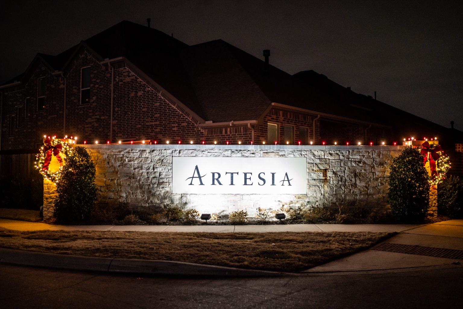 Artesia stone entrance monument with red and warm white bulbs along the top and illuminated wreaths with red bows at both ends.