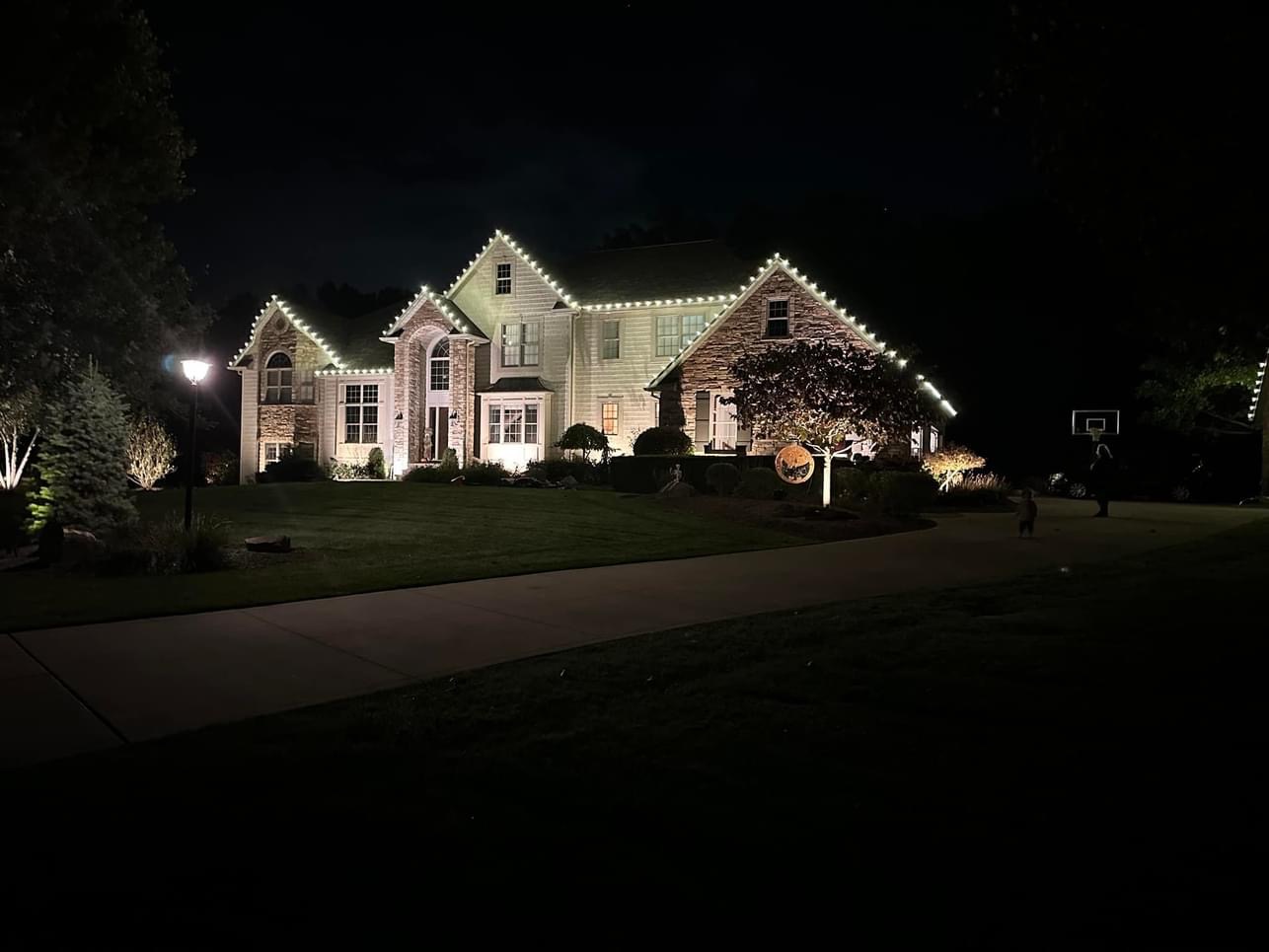 Warm white lights follow the roof peaks of a large home behind a curved driveway at night.