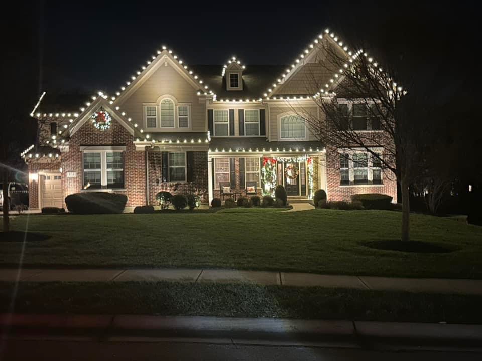 Warm white roofline bulbs frame a brick and siding house with an illuminated wreath and greenery around the entrance.