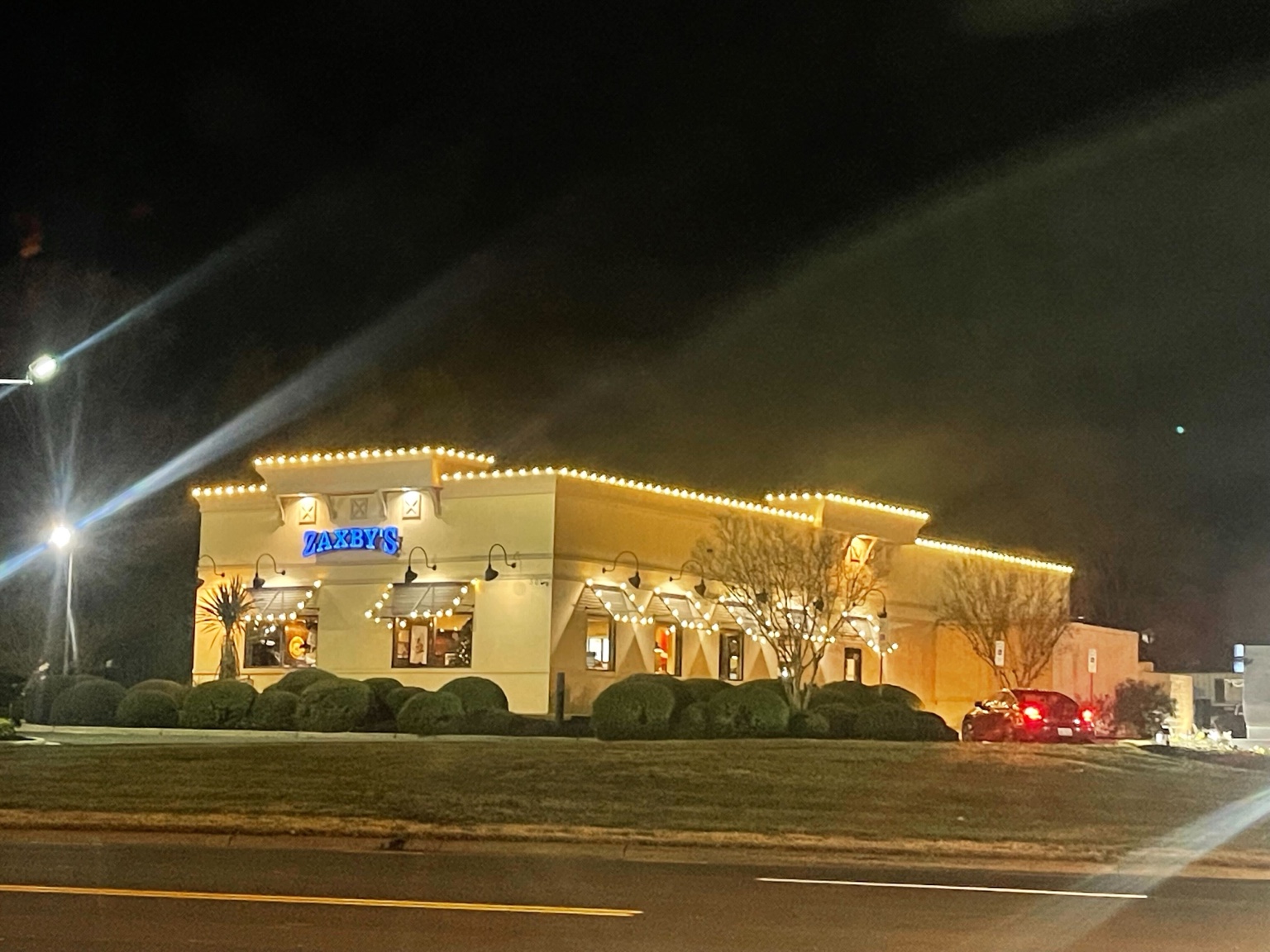 Warm white Christmas lights outline a Zaxby's restaurant roofline and window awnings at night.